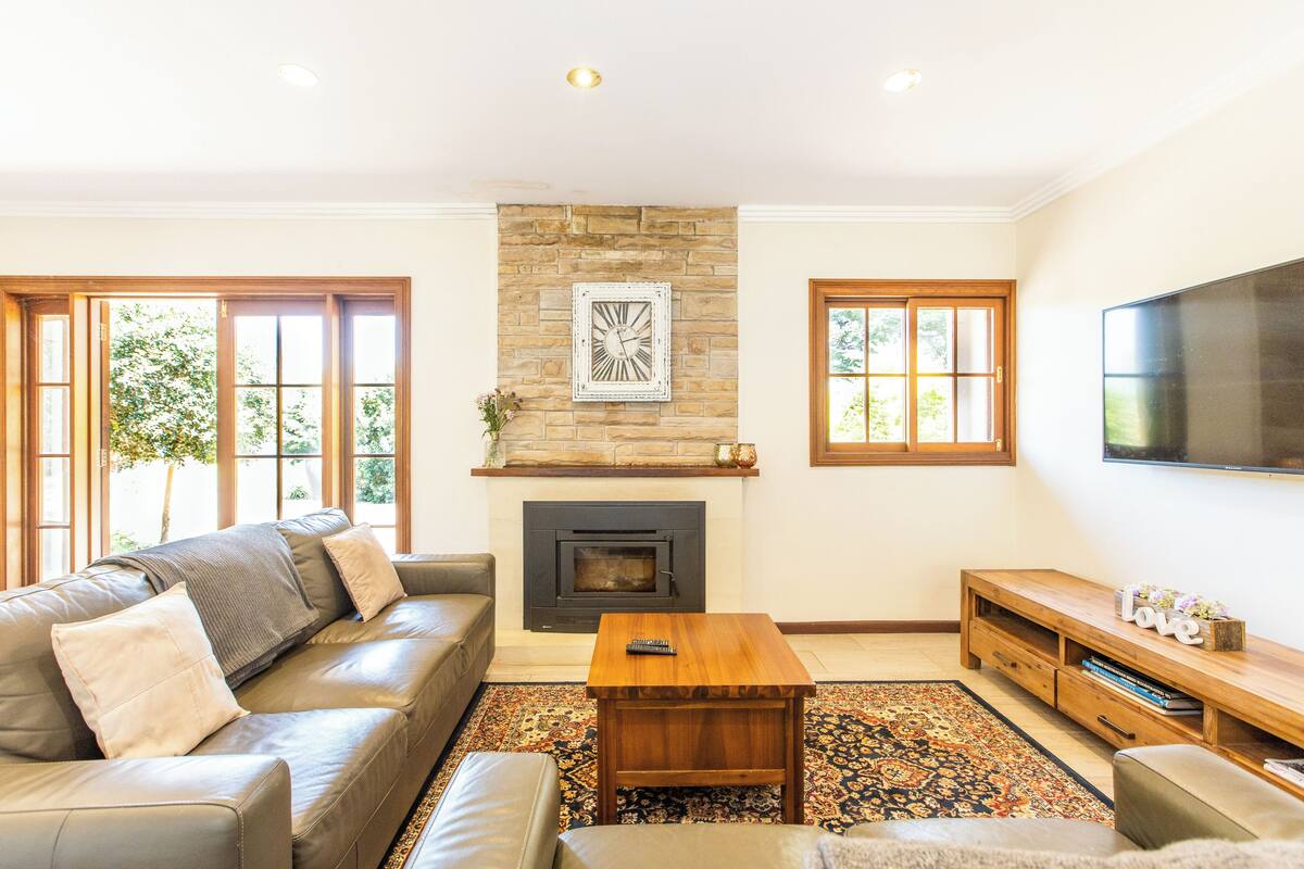 A living room with a stone fireplace, leather sofa, wooden coffee table, and a flat-screen TV mounted on the wall.