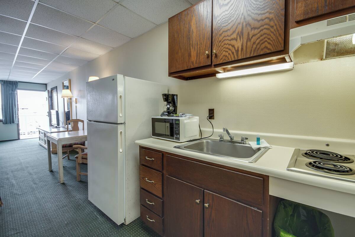 A compact kitchen with a refrigerator, microwave, sink, and stove. There is a dining table with chairs and a window with curtains.