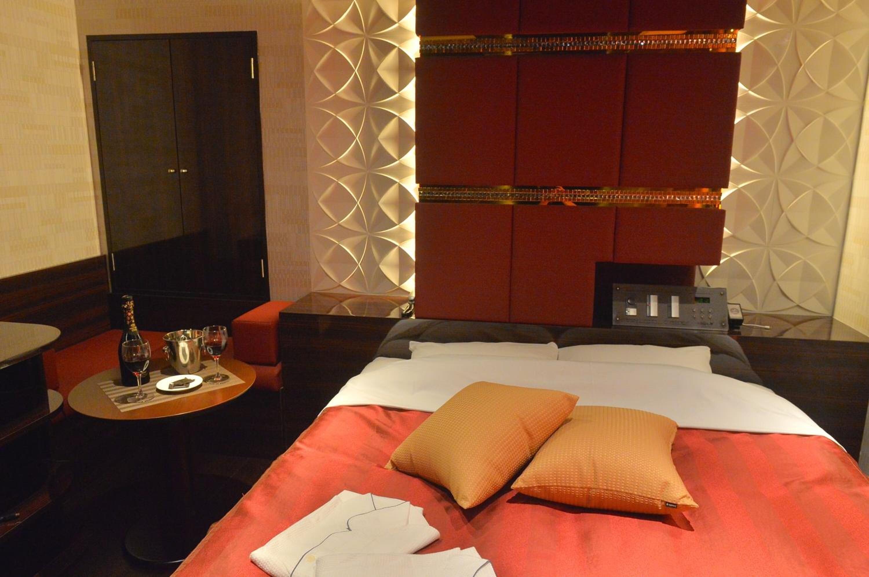 A hotel room with a bed, a bedside table with a bottle and glasses, a TV, and a wall with geometric patterns.