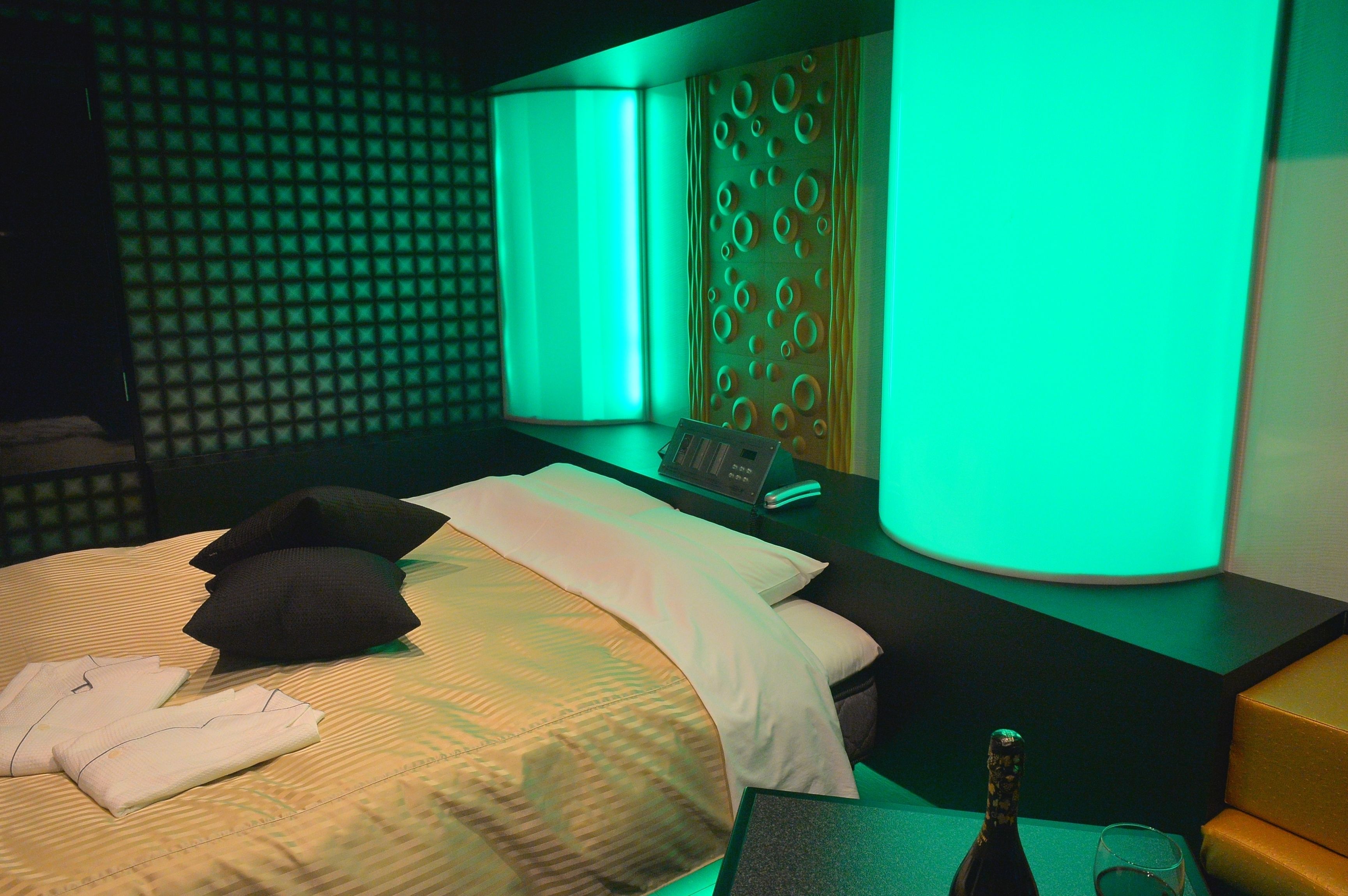 A hotel room with a bed, pillows, a bedside table with a bottle and glass, and a wall with a geometric pattern.