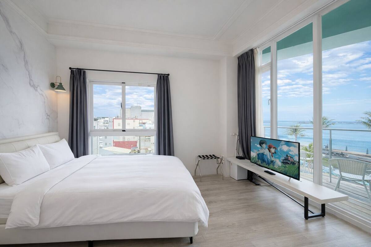 A hotel room with a large bed, a TV on a stand, a balcony with a view of the sea, and a cityscape visible through the window.