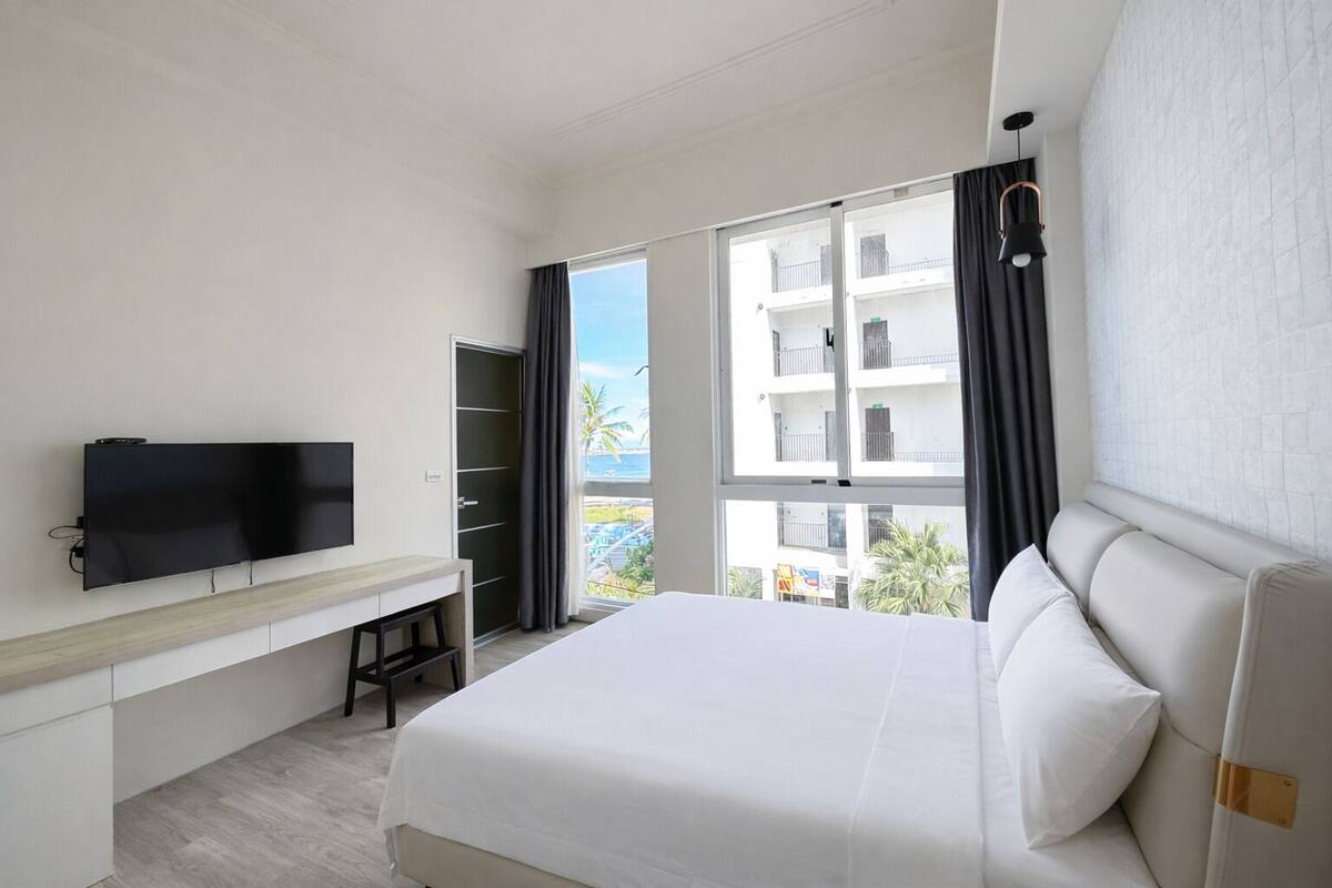 A hotel room with a bed, a desk, a TV, and a large window with a view of buildings and palm trees.