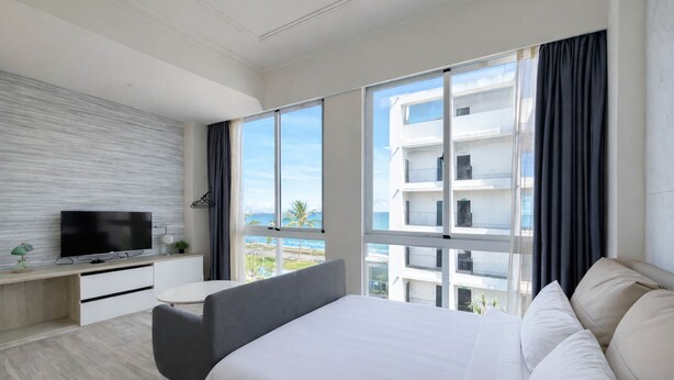 A modern hotel room with a large bed, a desk with a TV, and a view of a building and the sea. - Blue Ocean BnB (Hualien City)