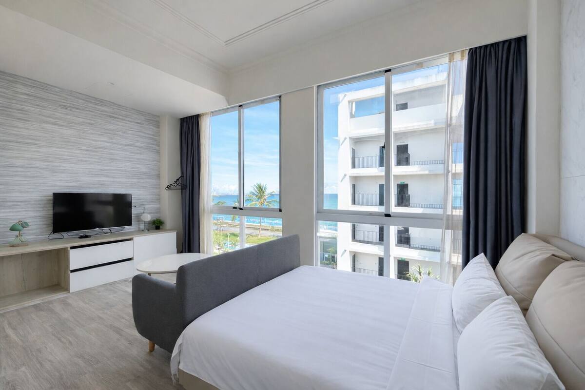 A modern hotel room with a large bed, a desk with a TV, and a view of a building and the sea.