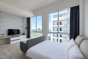 A modern hotel room with a large bed, a desk with a TV, and a view of a building and the sea. - Blue Ocean BnB (Hualien City)