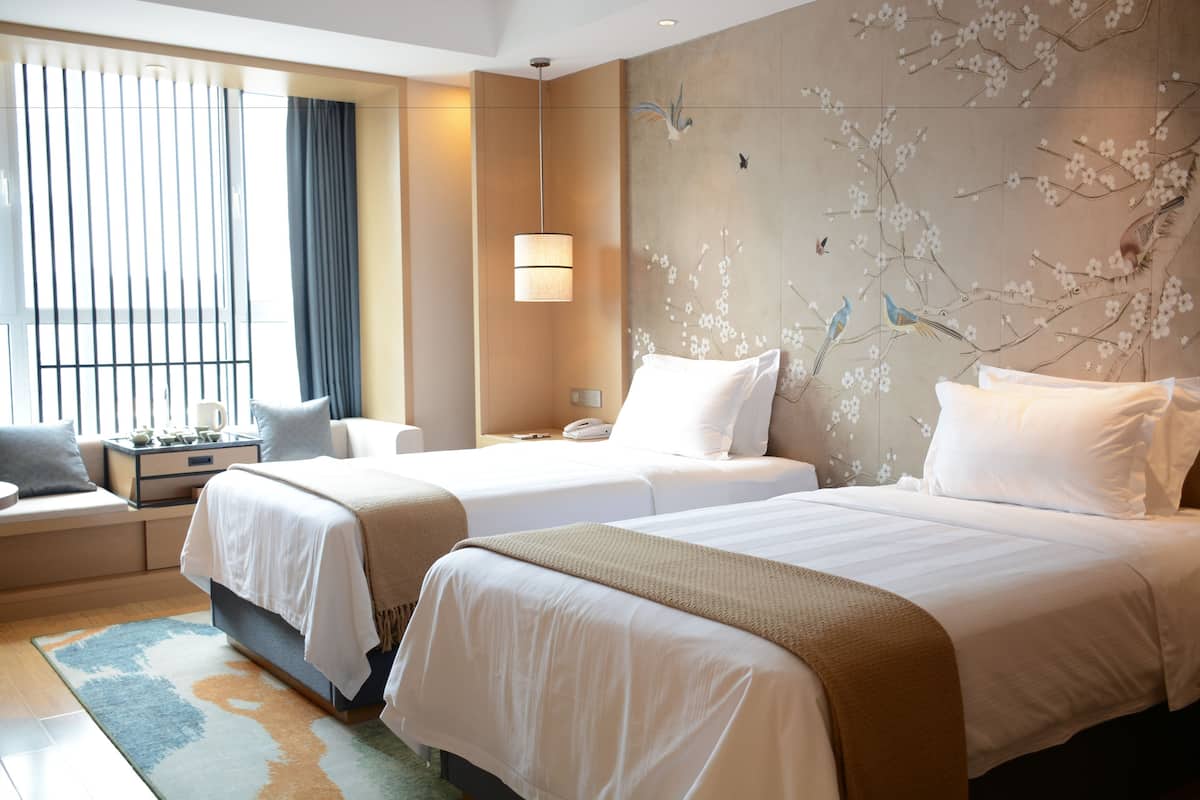 A hotel room with two beds, a sitting area with a sofa and armchair, a large window with blinds, a wall-mounted lamp, and a decorative wall with floral and bird designs.