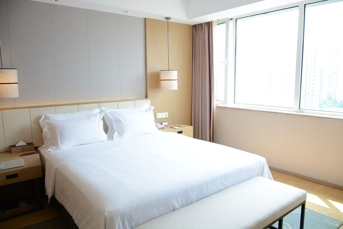 A hotel room with a large bed, bedside tables, a bench, a window with curtains, and a hanging light fixture.
