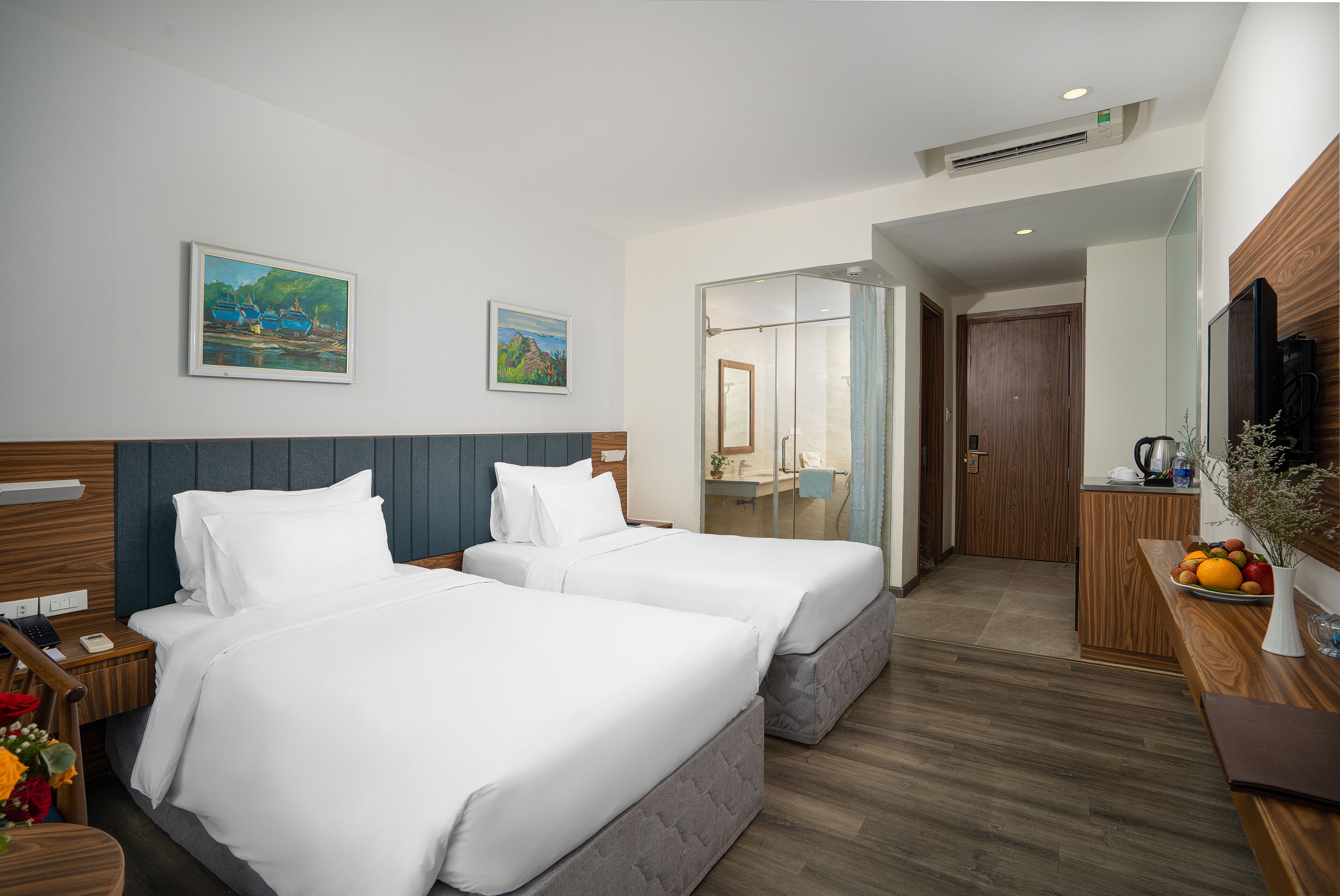 A hotel room with two beds, a wooden headboard, a desk with a vase of flowers, and a painting on the wall.