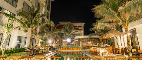 An outdoor dining area with tables set for guests, surrounded by palm trees and a pool.