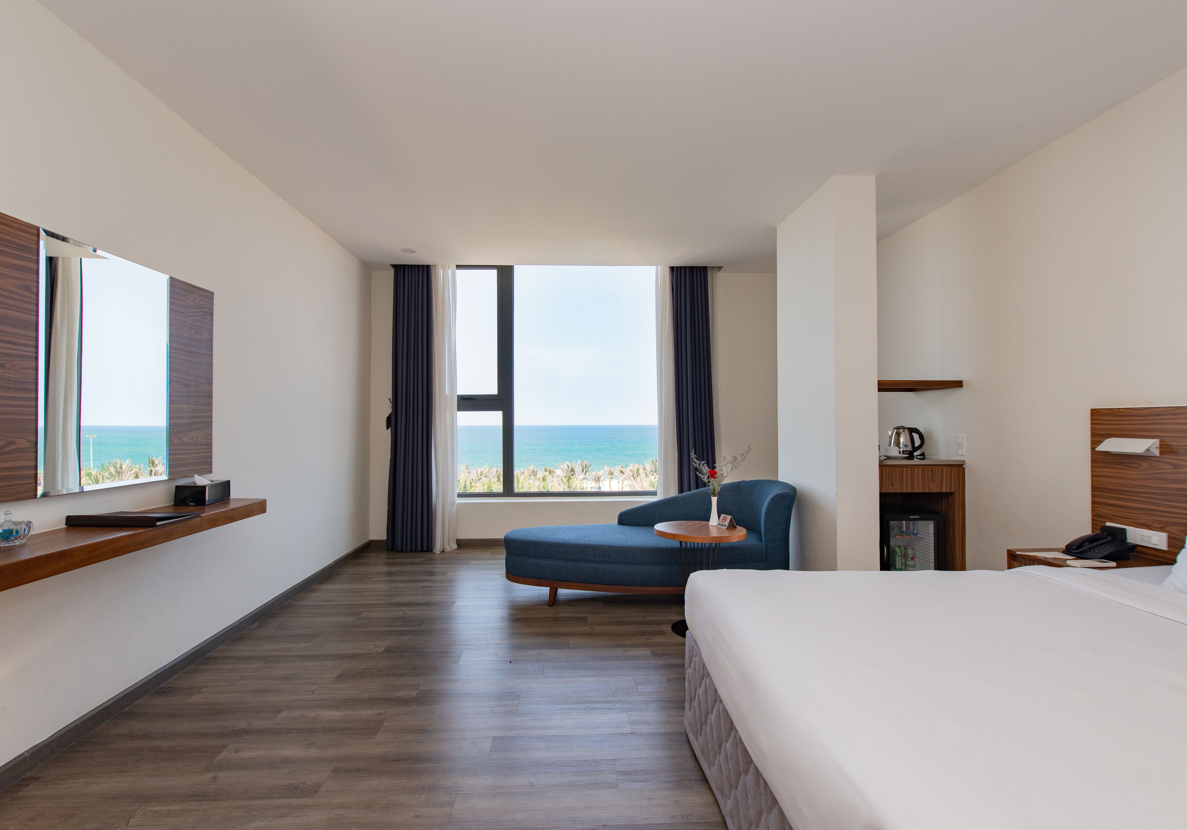 Habitación de hotel con una cama grande, un sofá, una mesita, una mesita de noche, un televisor y vista al océano.
