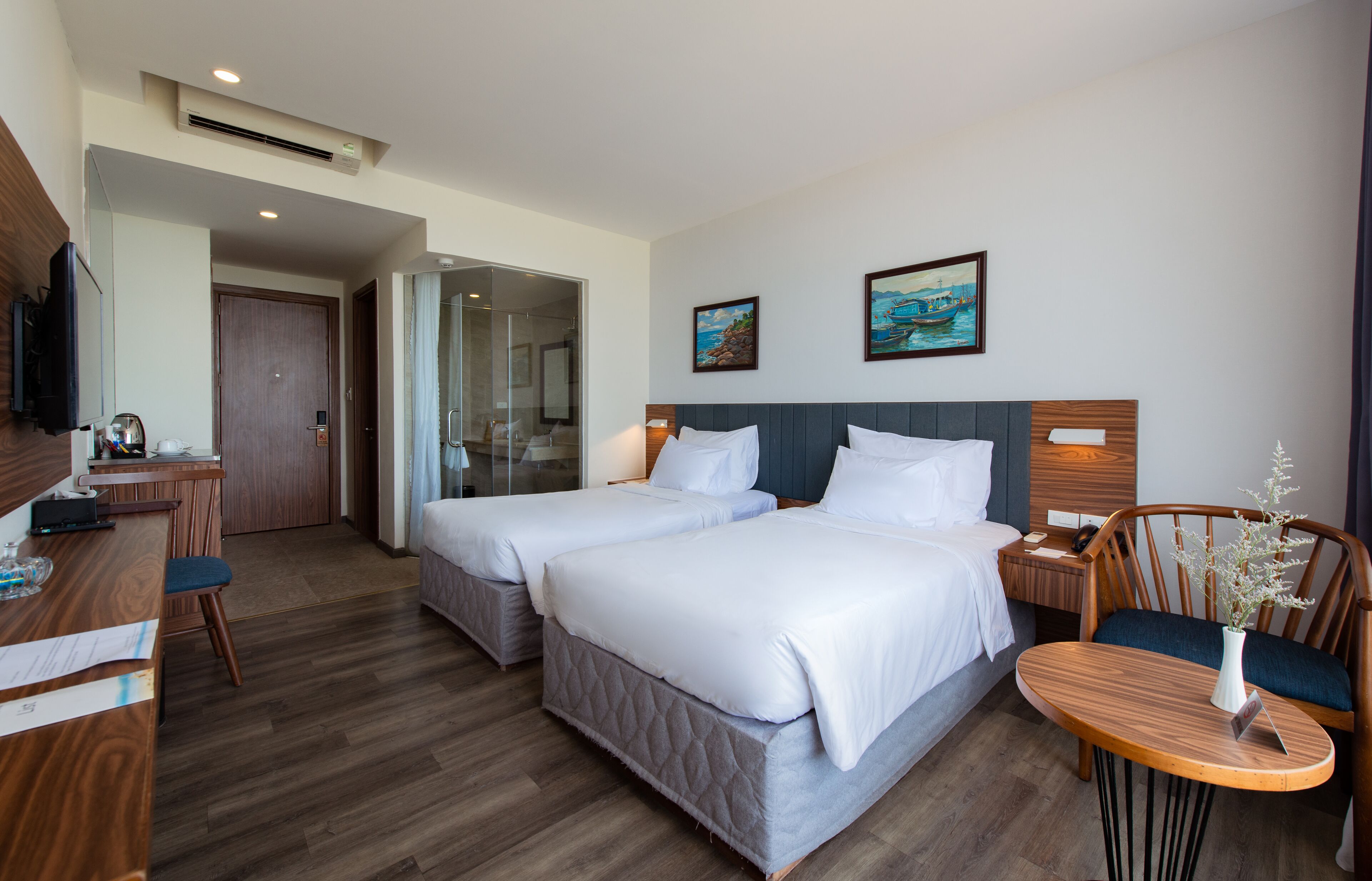 A hotel room with two beds, a desk, a chair, and a small table with a vase.