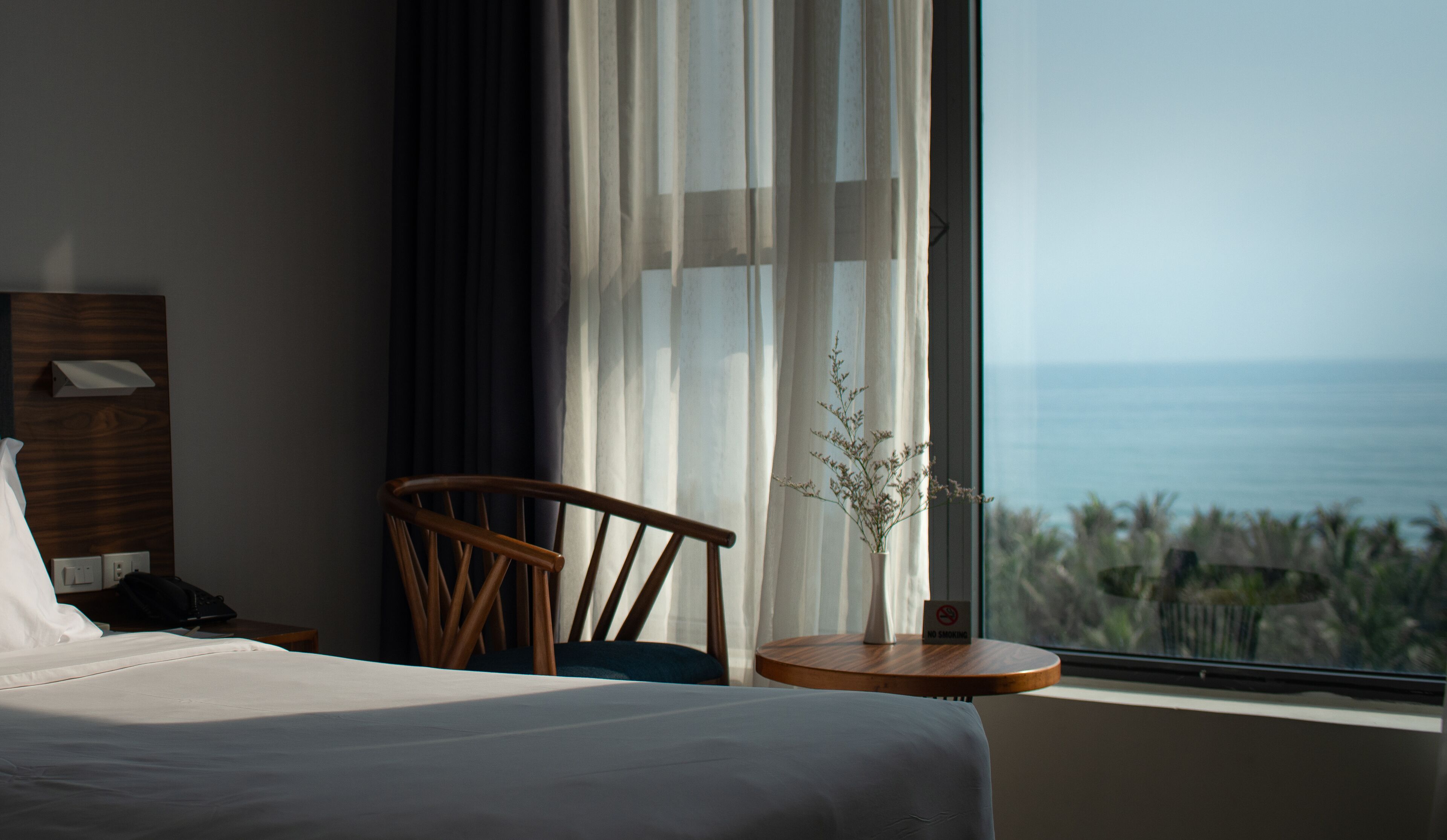 Habitación de hotel con cama, una silla, una mesita con un cartel de 'No Fumar' y vista al océano.