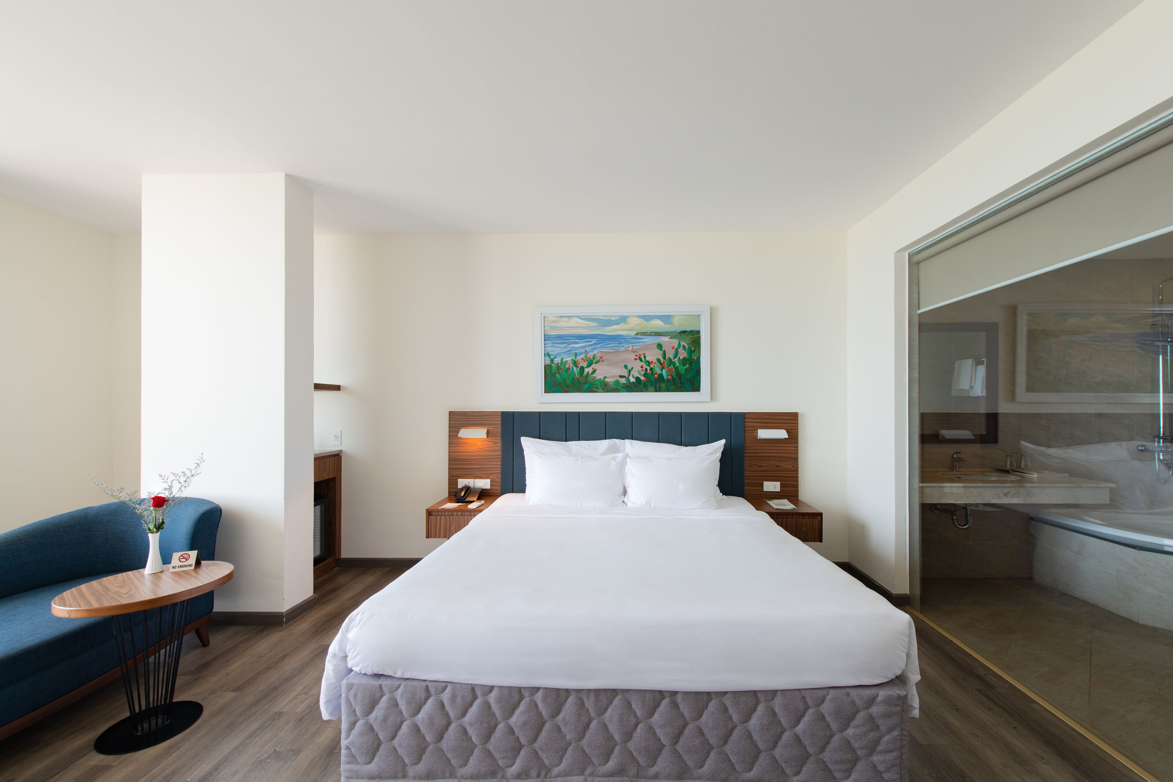 A hotel room with a bed, a blue sofa, a small table with a vase, and a painting on the wall.