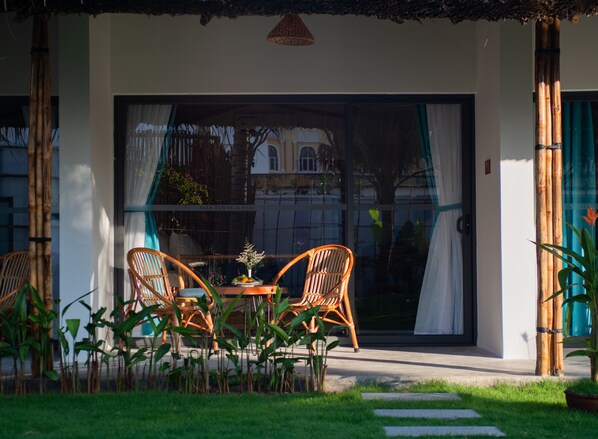 A patio with wicker chairs and a table, a glass door leading to a building, and a thatched roof overhead.