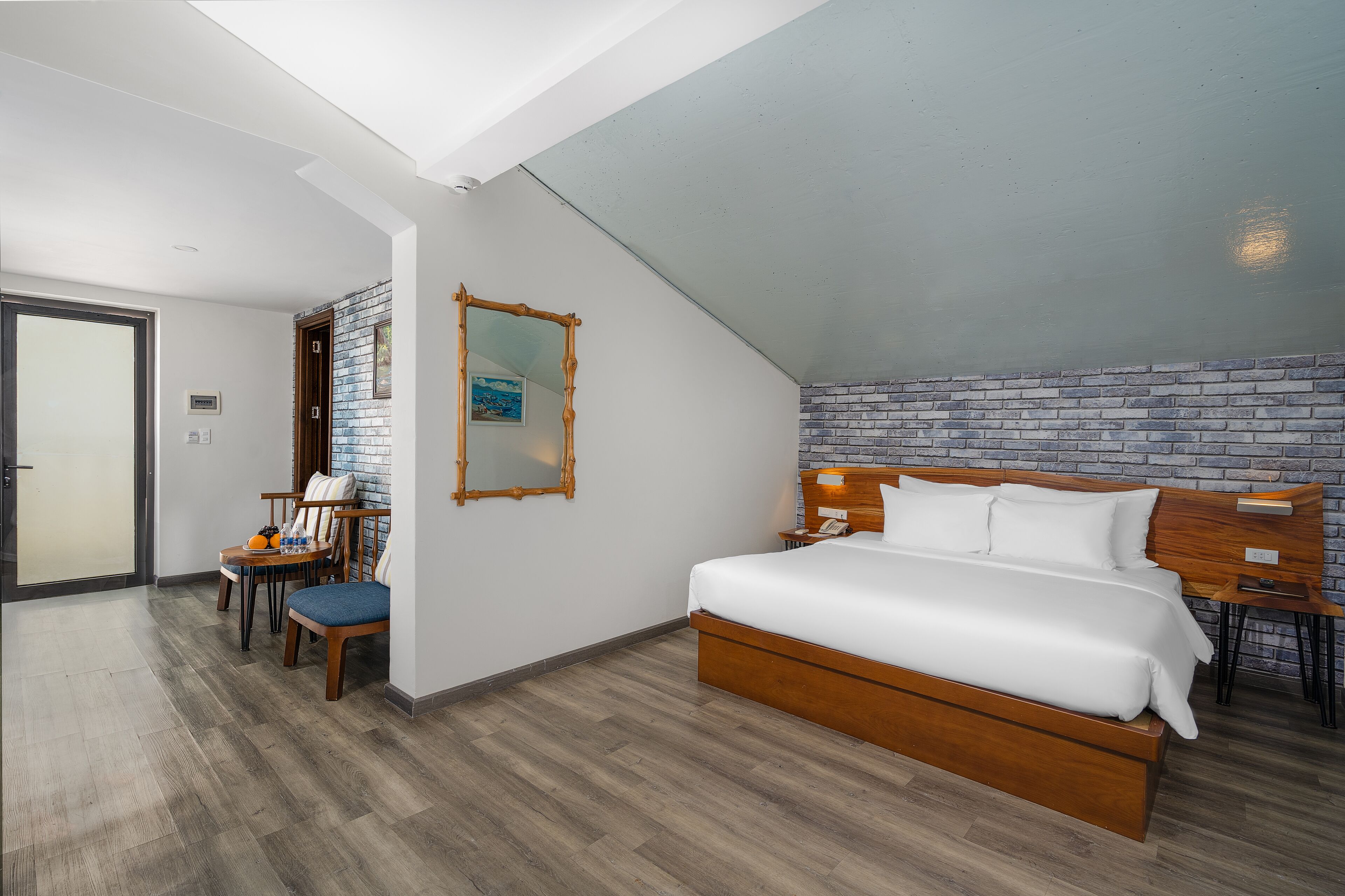 A bedroom with a wooden bed, a desk with chairs, a mirror, and a brick-patterned wall.