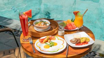 Une table avec des aliments pour le petit-déjeuner, notamment des œufs, du bacon et des toasts, placée près d’une piscine.