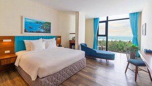 A hotel room with a bed, a desk, a chair, a painting, and a view of the ocean.