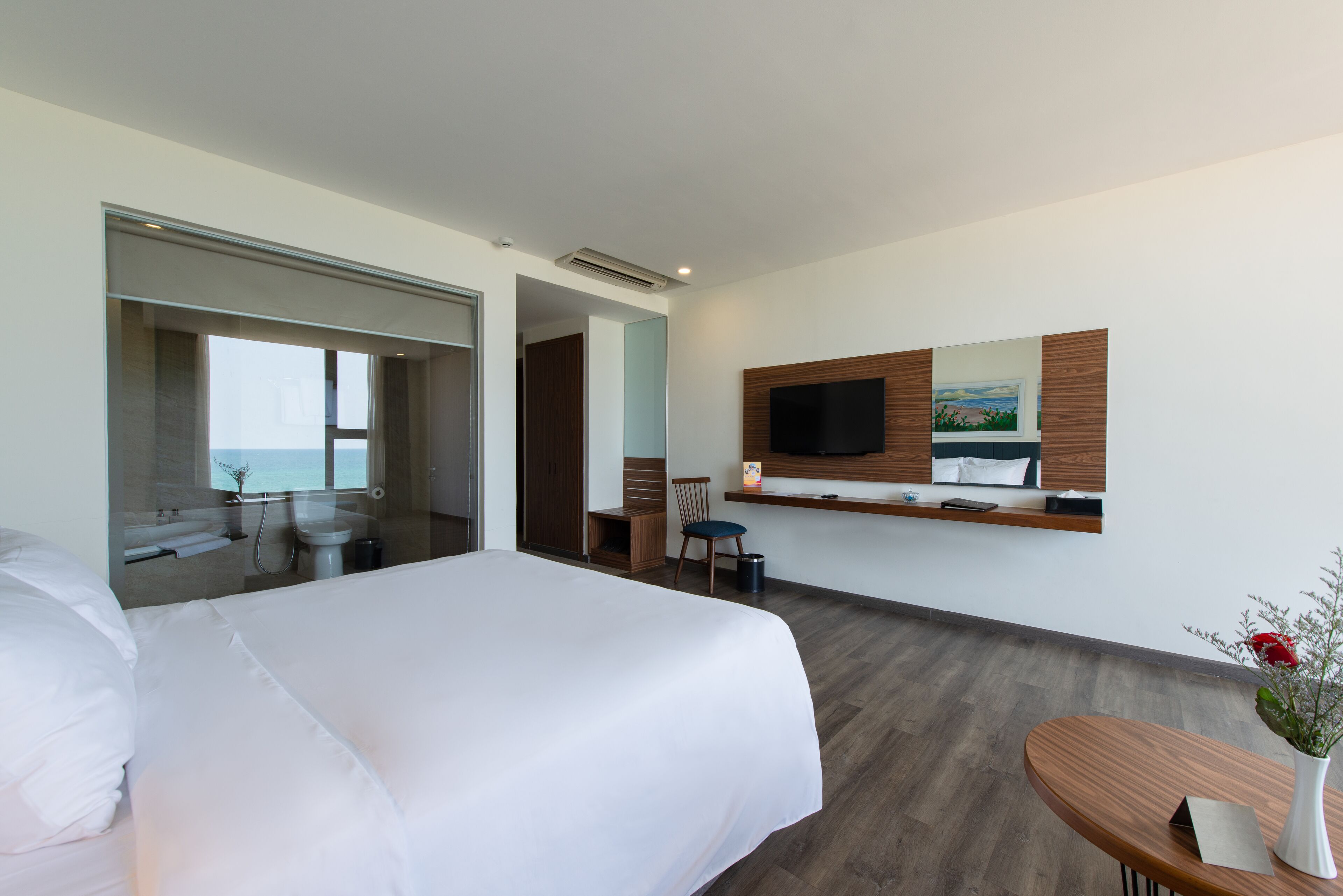 A modern hotel room with a large bed, a desk, a chair, a TV mounted on the wall, and a view of the sea through a glass door.
