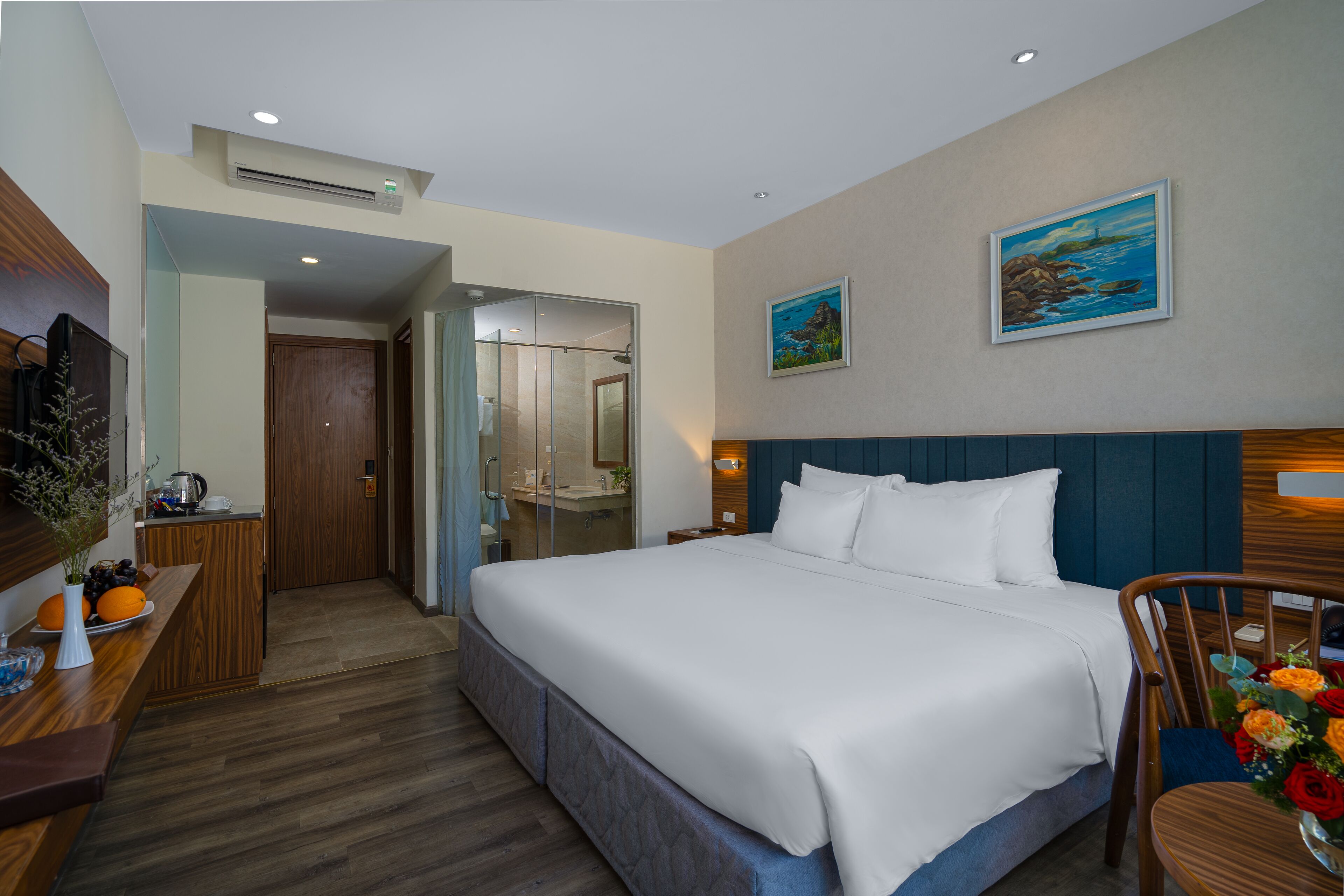 A hotel room with a large bed, wooden headboard, a desk with a vase of flowers, and a painting on the wall.