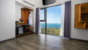 A room with a wooden floor, a TV mounted on the wall, a wardrobe, and a large window with a view of the sea.