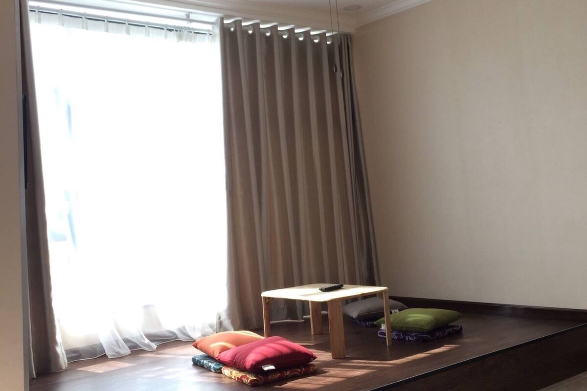 Une pièce avec un ventilateur de plafond, une fenêtre avec des rideaux, une petite table avec une télécommande et un banc avec des coussins.