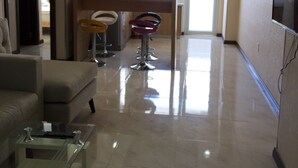 A modern living room with a glass coffee table, a sofa, and a TV mounted on the wall. There is a kitchen area with a bar stool and pendant lights. - Hoang Ky Hotel (Bac Nha Trang)