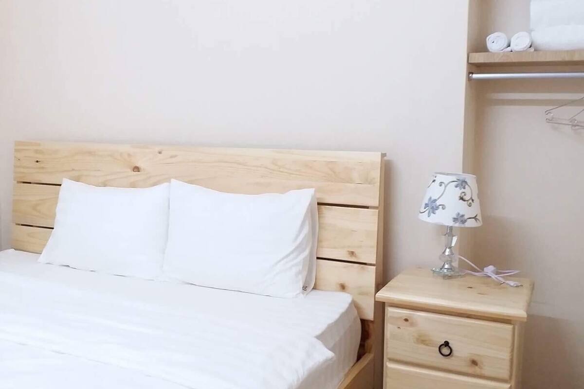 Un lit en bois avec une literie blanche, une table de chevet avec une lampe et une étagère avec des serviettes.