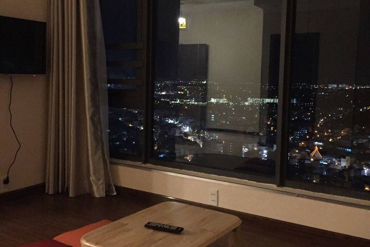 Une pièce avec une grande fenêtre donnant sur une vue de la ville la nuit, une table en bois et une télécommande posée dessus.