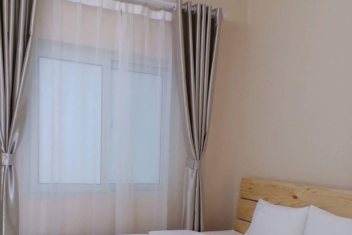 Un lit bien fait, avec du linge de lit blanc, dans une chambre dotée d’une tête de lit en bois et d’une fenêtre ornée de rideaux légers.