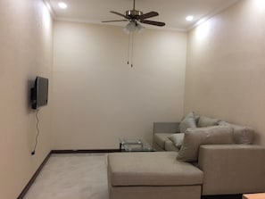 A room with a ceiling fan, a flat-screen TV mounted on the wall, a grey sofa with cushions, and a glass coffee table.