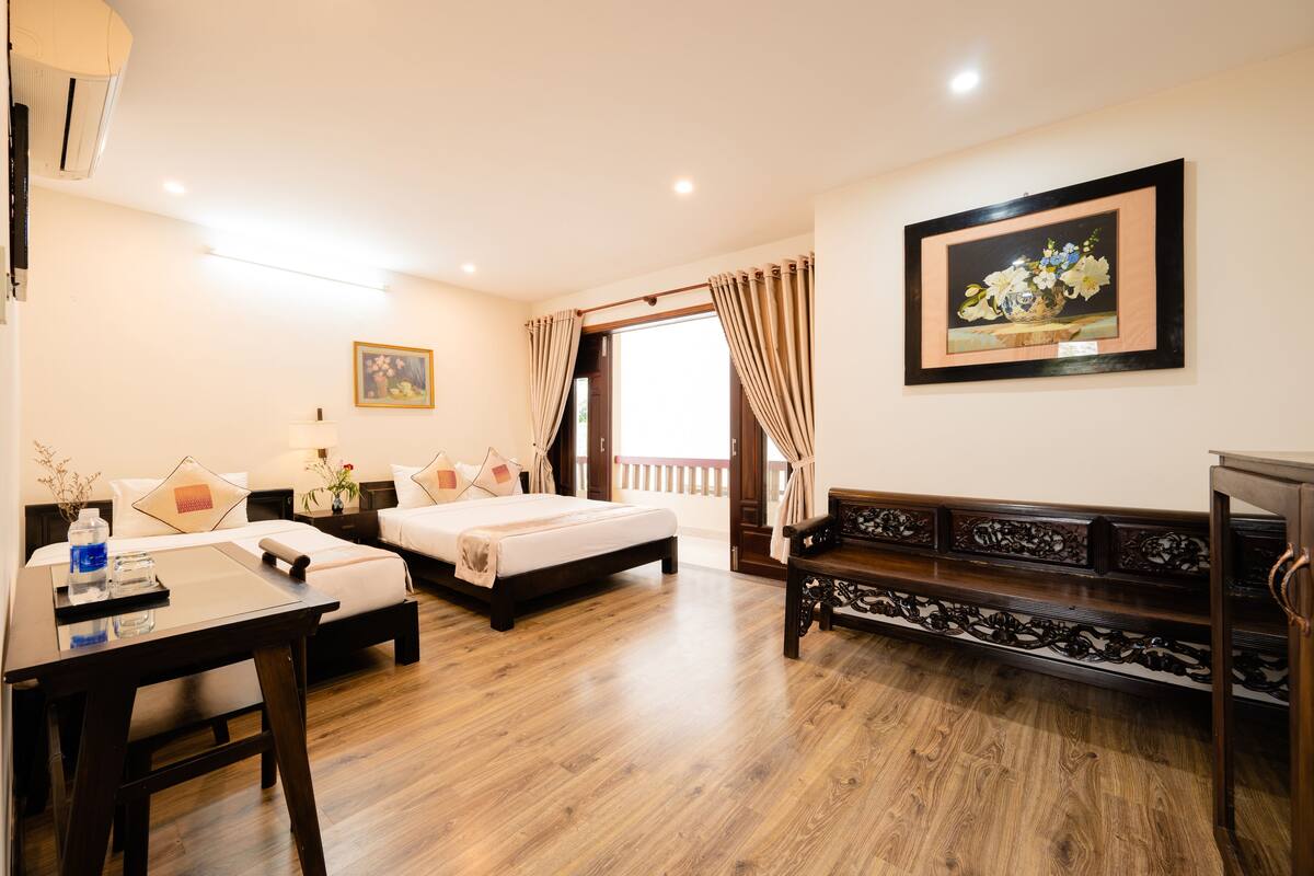 A hotel room with a wooden floor, two beds, a desk, a chair, a bench, a painting, and a window with curtains.