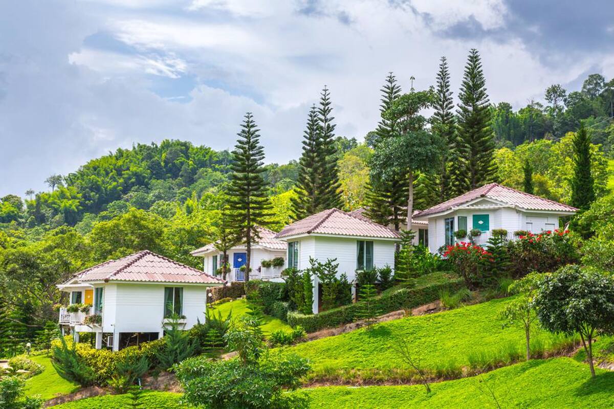 A row of white cottages with red roofs, surrounded by lush greenery and tall trees, on a hillside.