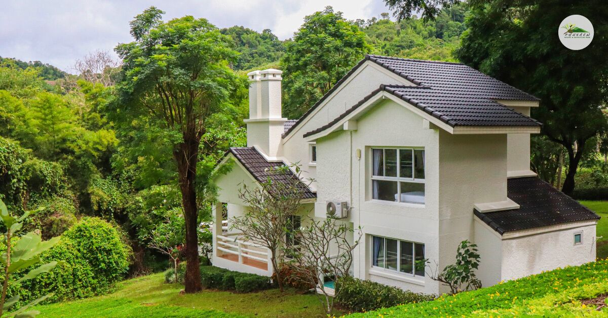 Een huis met twee verdiepingen, een witte gevel en donkere dakpannen, omringd door weelderige begroeiing en bomen.