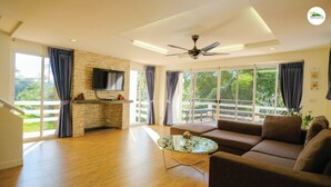 A spacious living room with a stone fireplace, a flat-screen TV, a ceiling fan, and large windows offering a view of greenery.