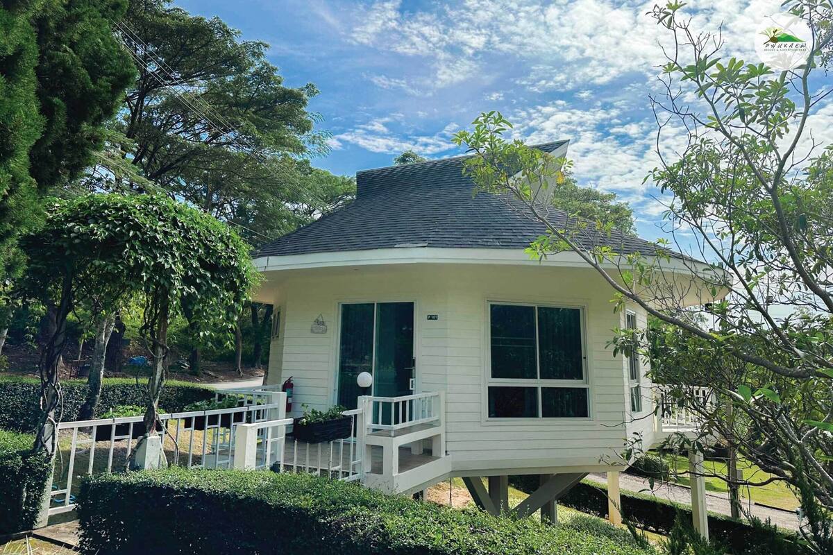 A small, single-story house with a balcony, surrounded by lush greenery and trees.