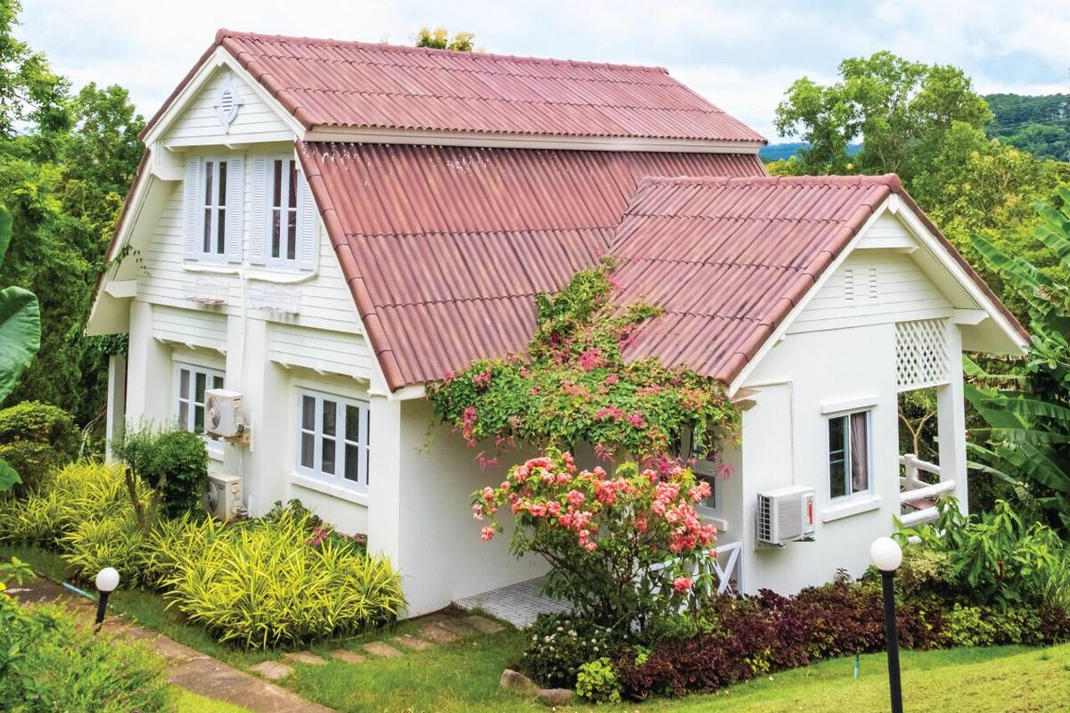 A white house with a red roof, surrounded by lush greenery and flowering plants.