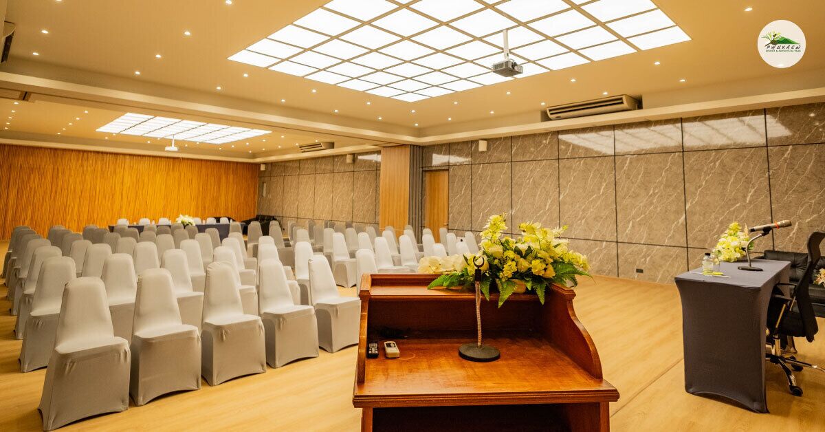 Une salle de conférence avec des rangées de chaises blanches, un pupitre équipé d’un microphone et un arrangement floral.
