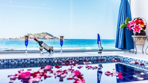 A pool area with a view of the sea and a mountain, a bottle of champagne, and a vase with flowers.
