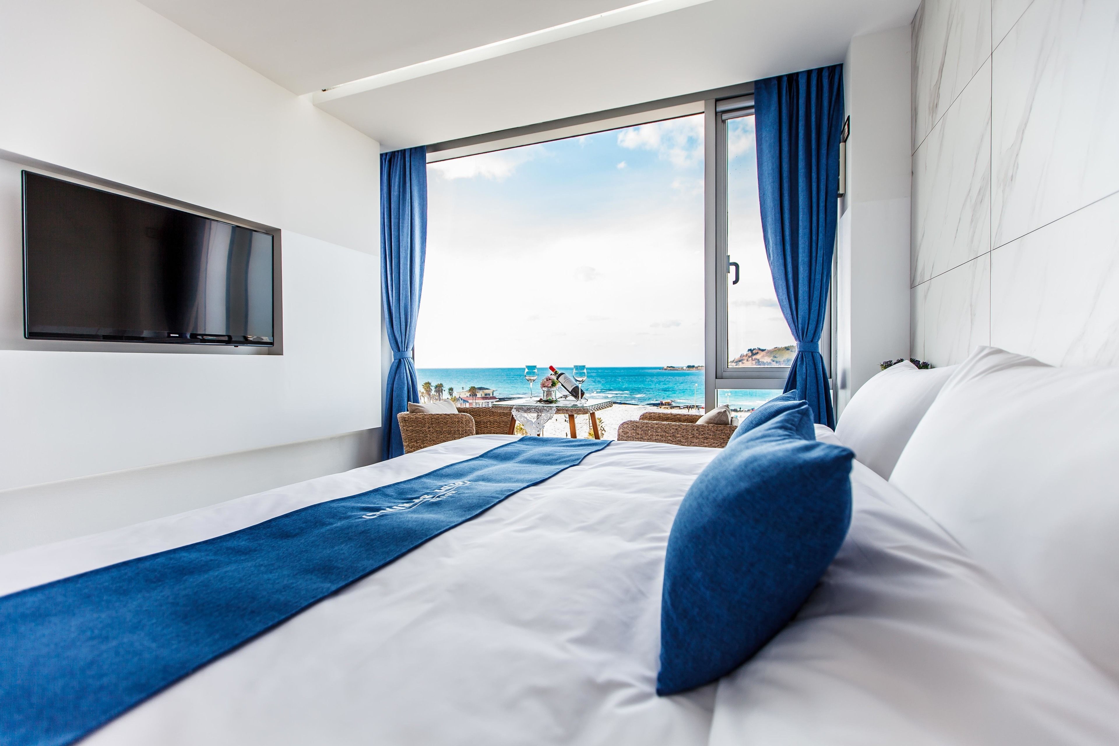 Habitación de hotel con una cama grande, un televisor montado en la pared y vistas a la playa a través de un amplio ventanal.