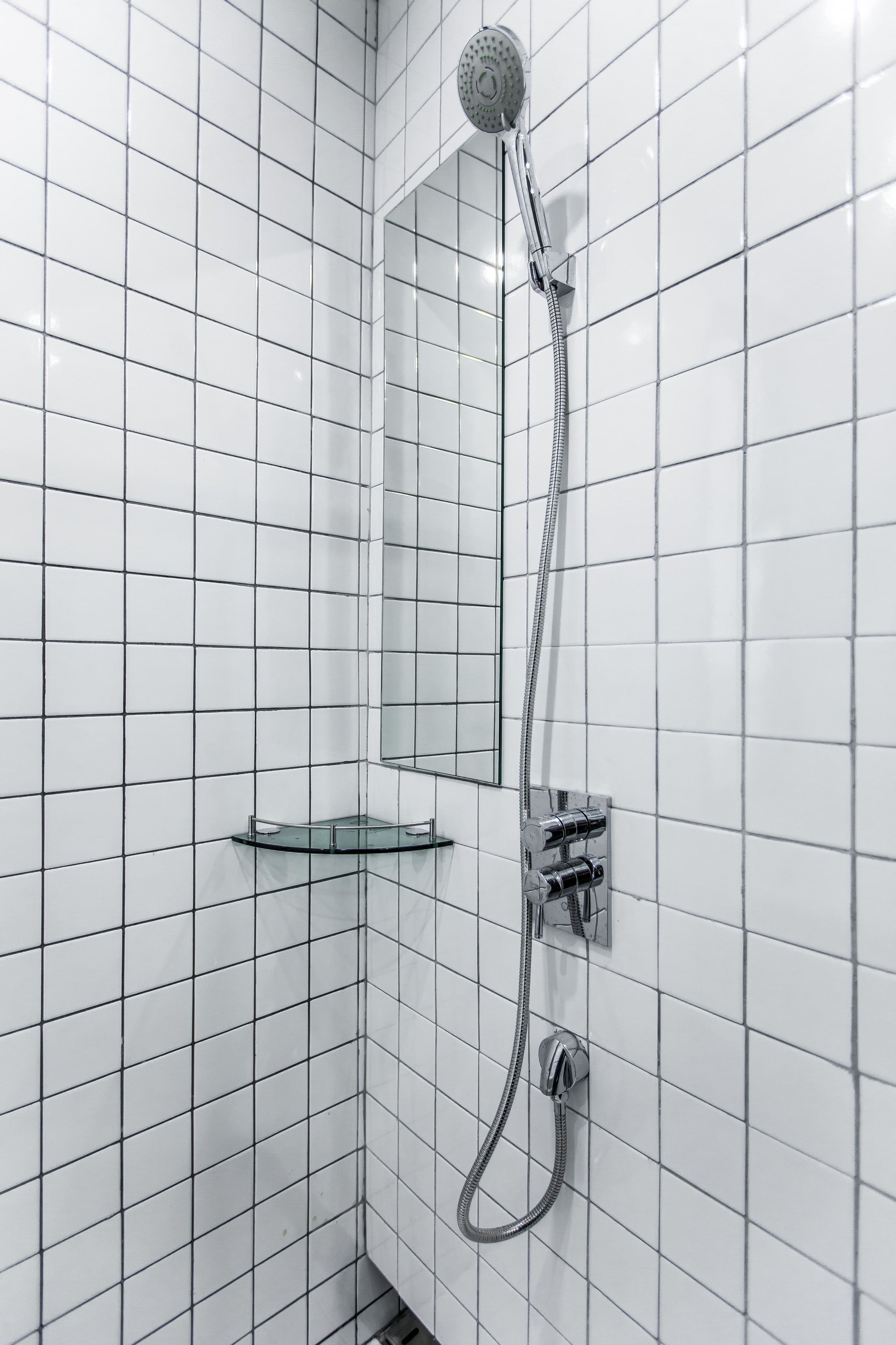 Une douche d’angle avec un pommeau de douche à main, une étagère en verre et un panneau de commande fixé au mur.