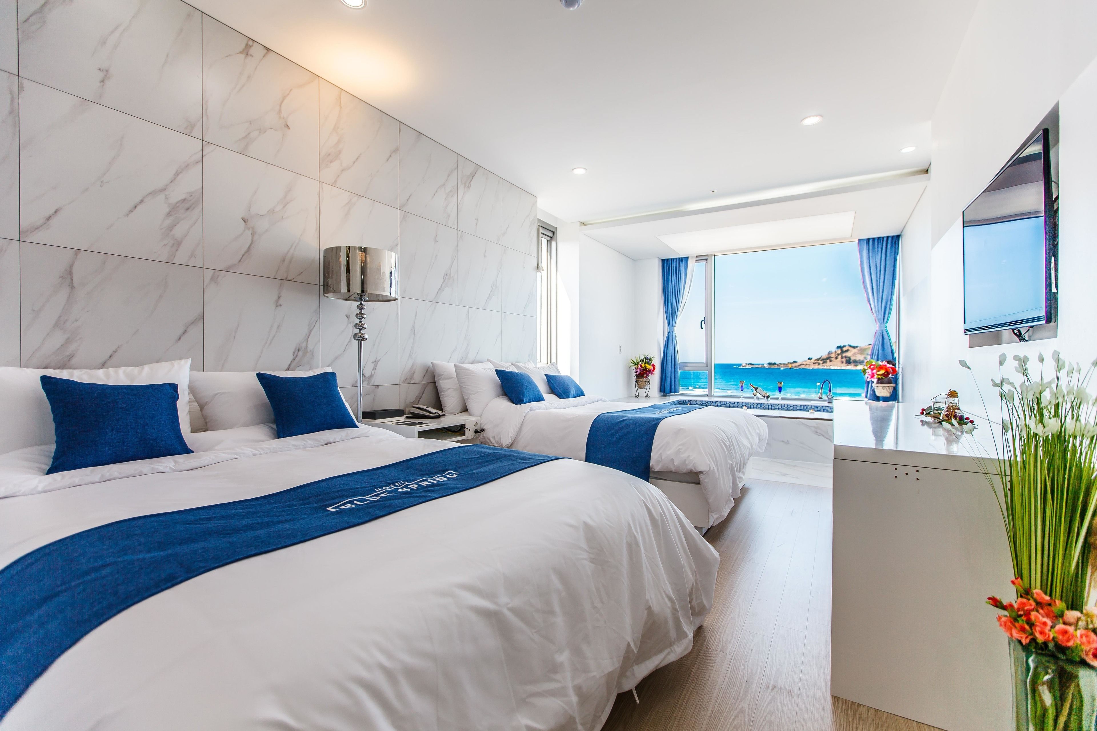 Una habitación de hotel moderna con dos camas, paredes de mármol, un ventanal con vista al mar y un televisor de pantalla plana.