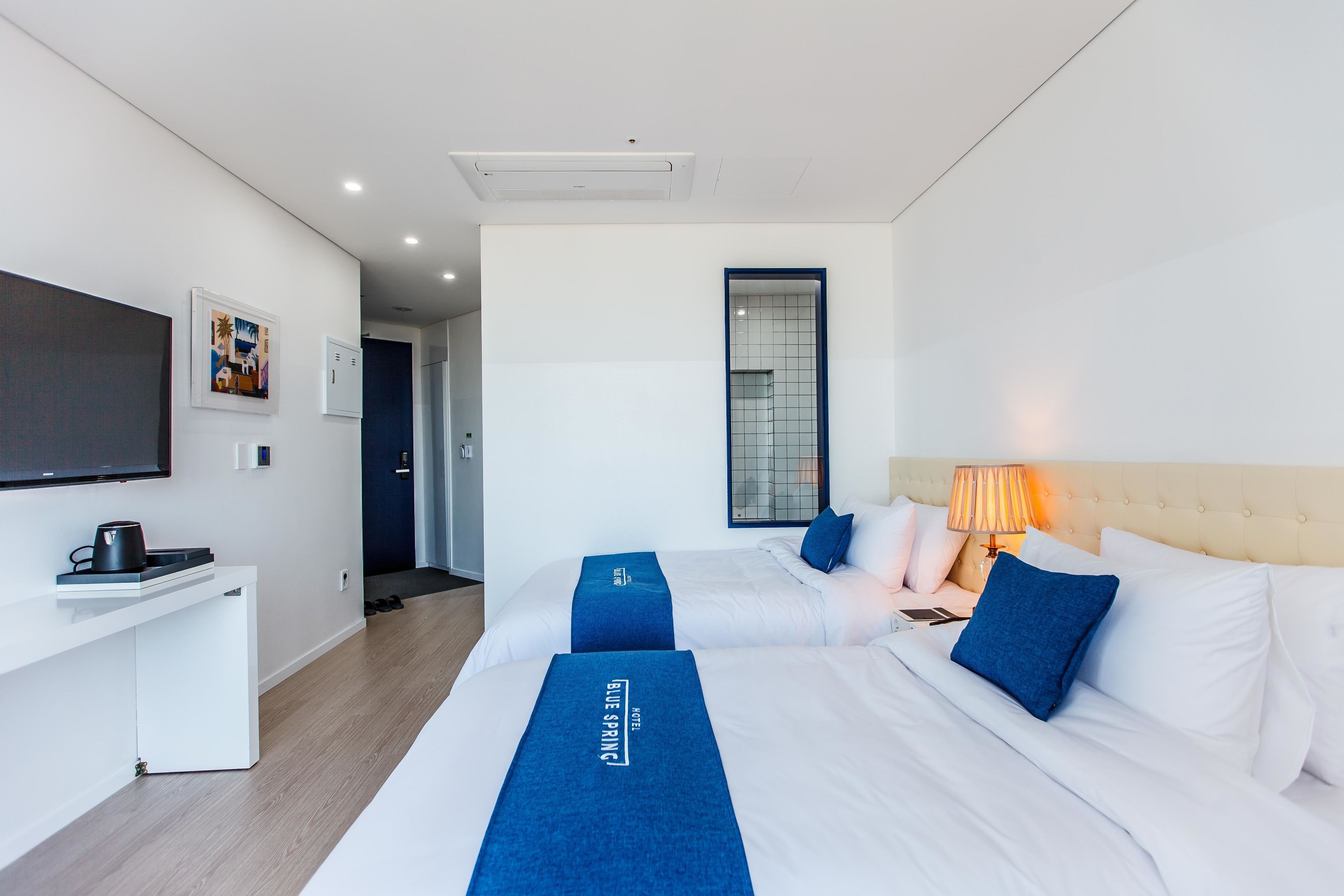 A hotel room with two beds, a desk, a TV, and a bathroom visible in the background.