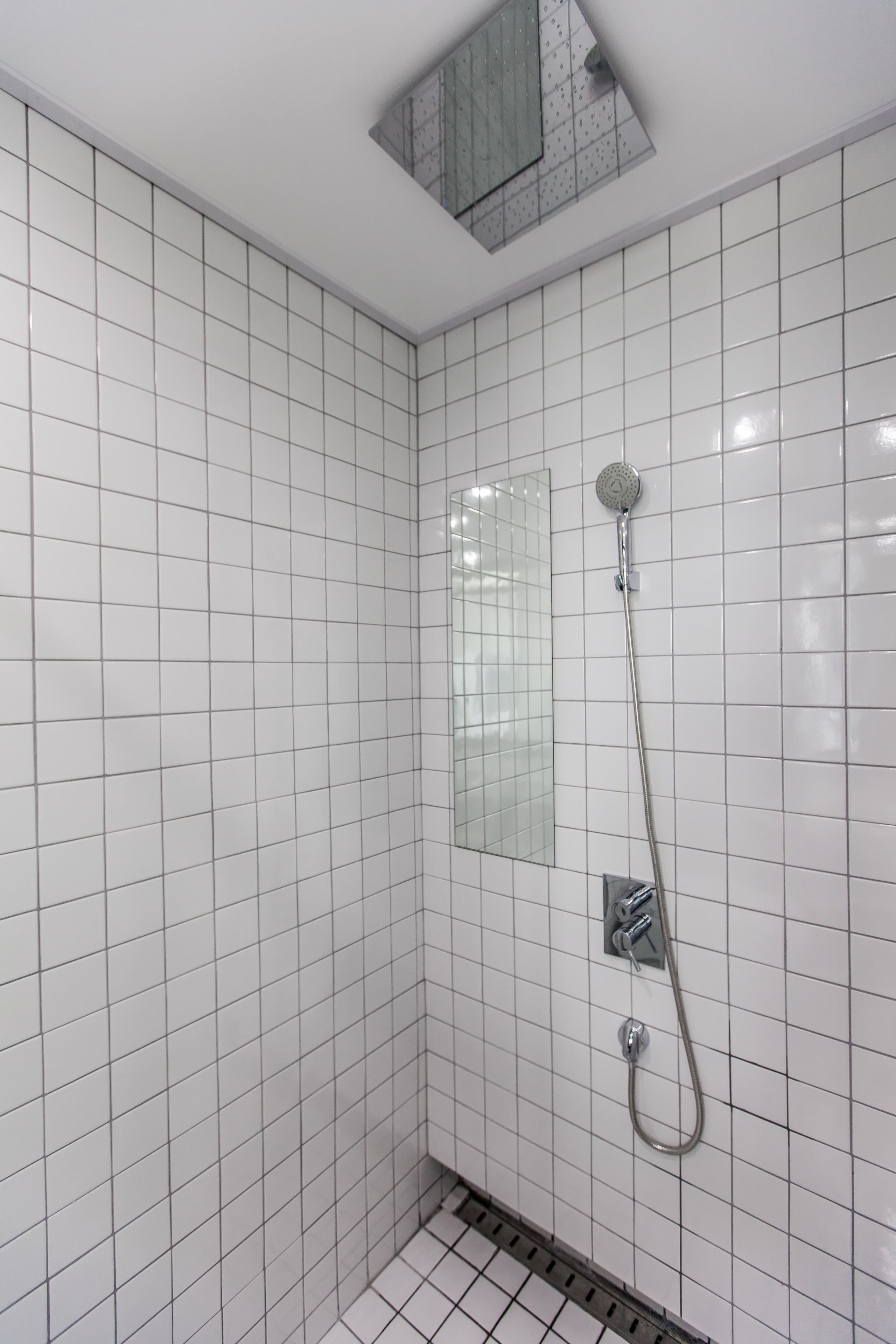 A bathroom with white tiled walls and floor, a shower with a handheld showerhead, and a small window with a grid pattern.