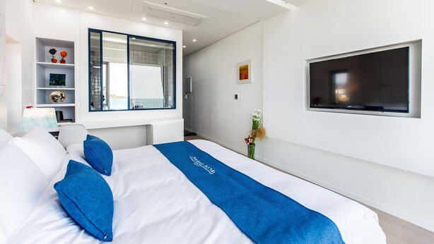 A modern hotel room with a large bed, a TV mounted on the wall, a built-in wardrobe, and a small desk with a lamp. - Bluespring Boutique Hotel (Jeju City)