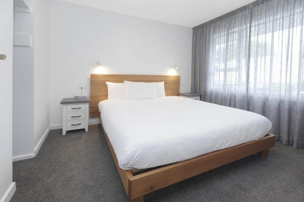 A modern hotel room with a wooden bed, white bedding, a bedside table with a lamp, and a large window with curtains.