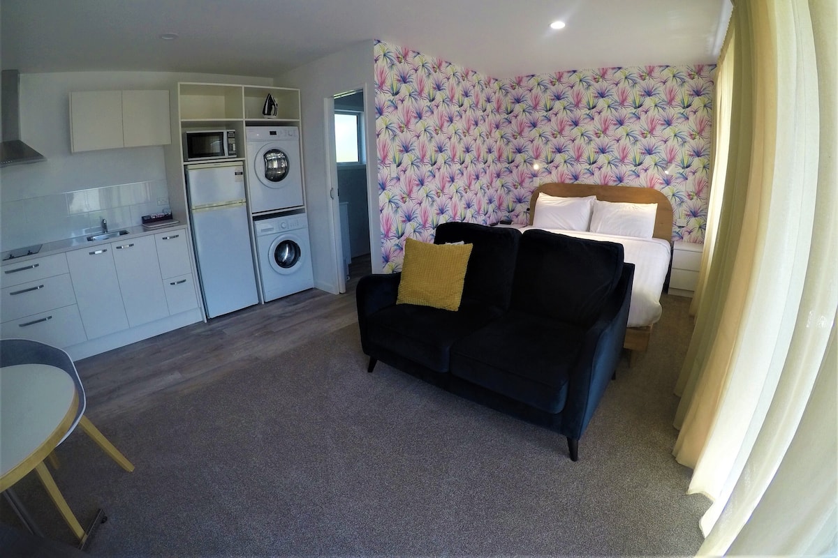 A compact hotel room with a kitchenette, a bed with white linens, a black sofa with a yellow pillow, and a patterned wallpaper.