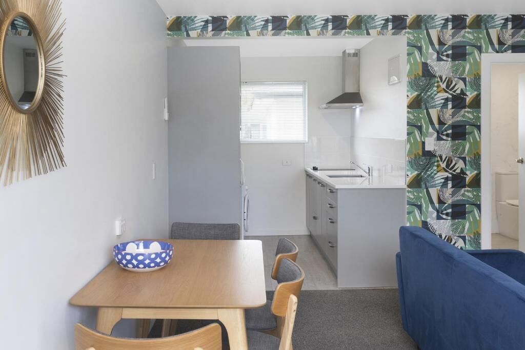 Une cuisine compacte avec un plan de travail blanc, un évier et une cuisinière. On y trouve également un coin repas avec une table et des chaises en bois.
