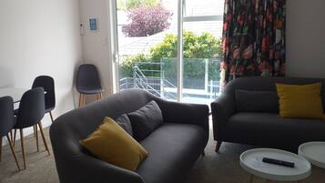 A modern living room with a grey sofa, a smaller grey armchair, and a white coffee table. There is a window with a view of a balcony and greenery outside.