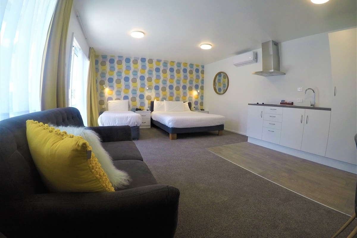 A room with two beds, a sofa, a kitchen area, and a patterned wall.
