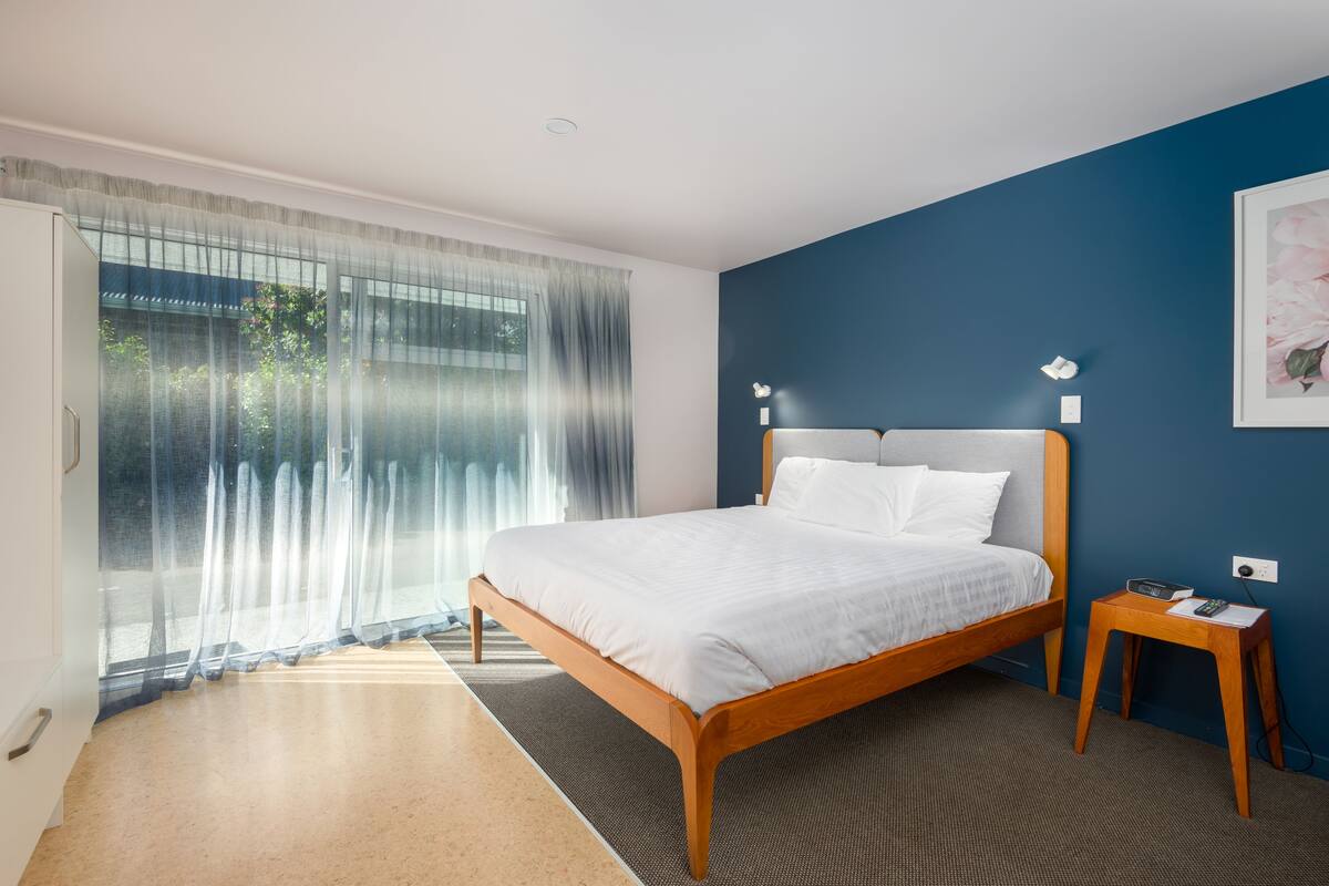 A modern bedroom with a wooden bed, a nightstand, and a large window with sheer curtains.